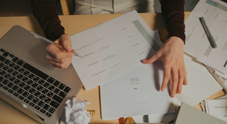 Pessoa analisando documentos e planilhas ao lado de um notebook, representando planejamento para comprar ou alugar um imóvel.